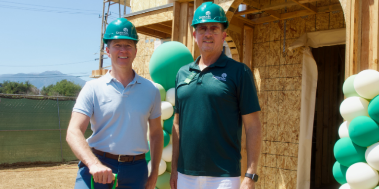Two construction professionals wearing Genesis Builders hard hats stand in front of a wood-framed home under construction, with green and white balloon decorations at the building site entrance.